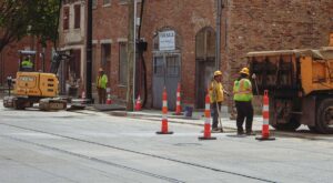 In the foreground, construction workers wearing safety hats and vests stand on a road surrounded by safety cones and a construction dump truck.